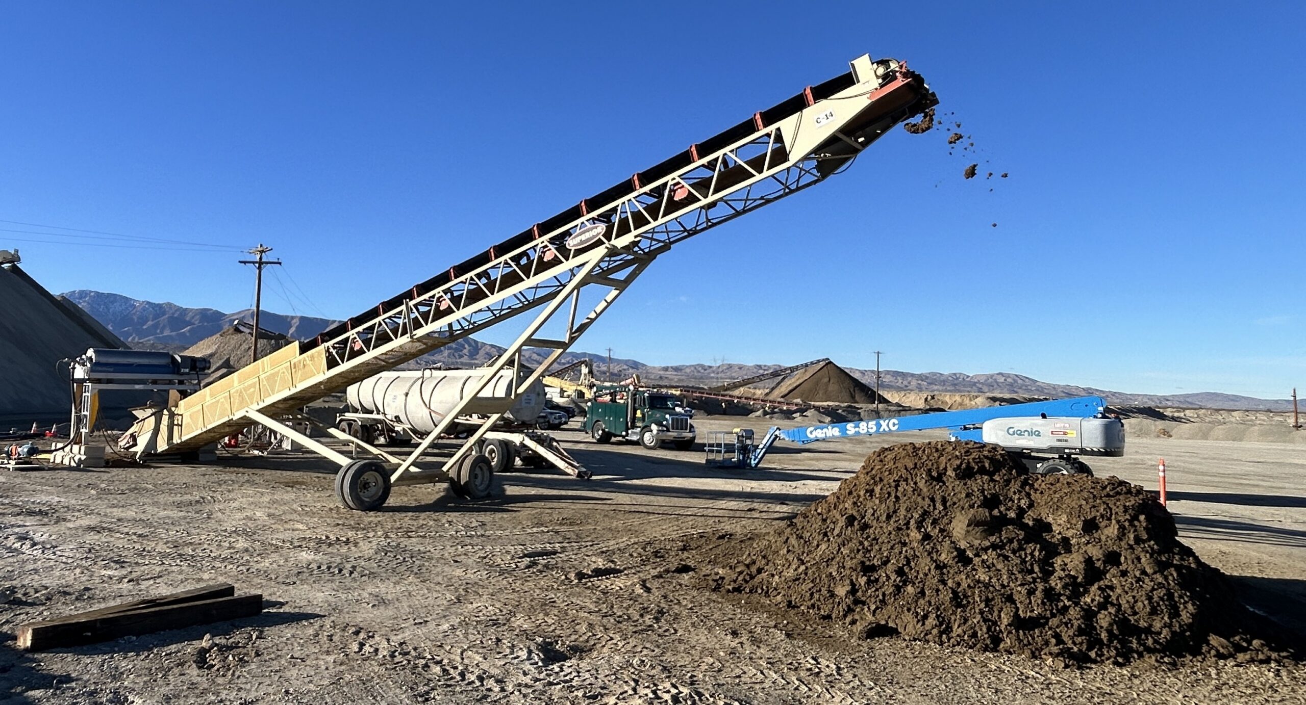 A large belt conveyor with a pile of dirt below it. The machinery is outside with blue skies behind it.