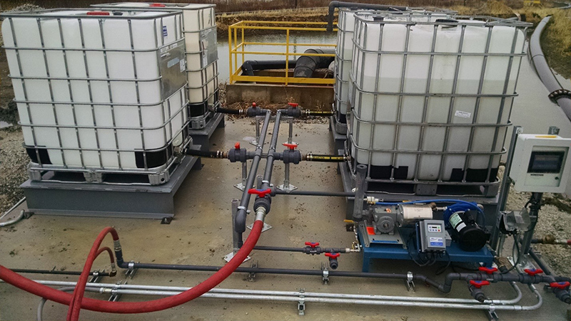 Quatre conteneurs de polymère liquide reliés à un patin de dosage de polymère liquide installé à côté d’un bassin de décantation.