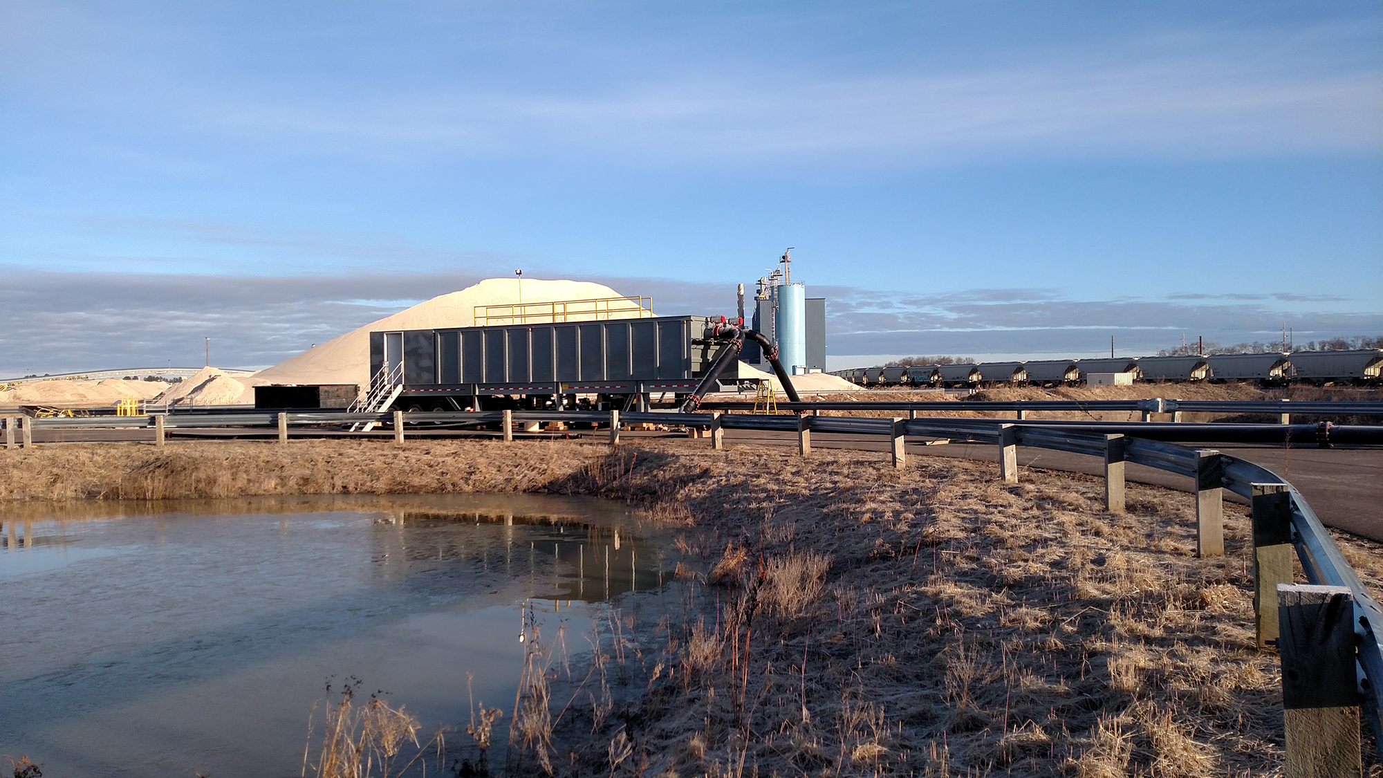 A rectangular water clarifier setup next to a stormwater holding pond with sand mounds and a train on each side of the background.