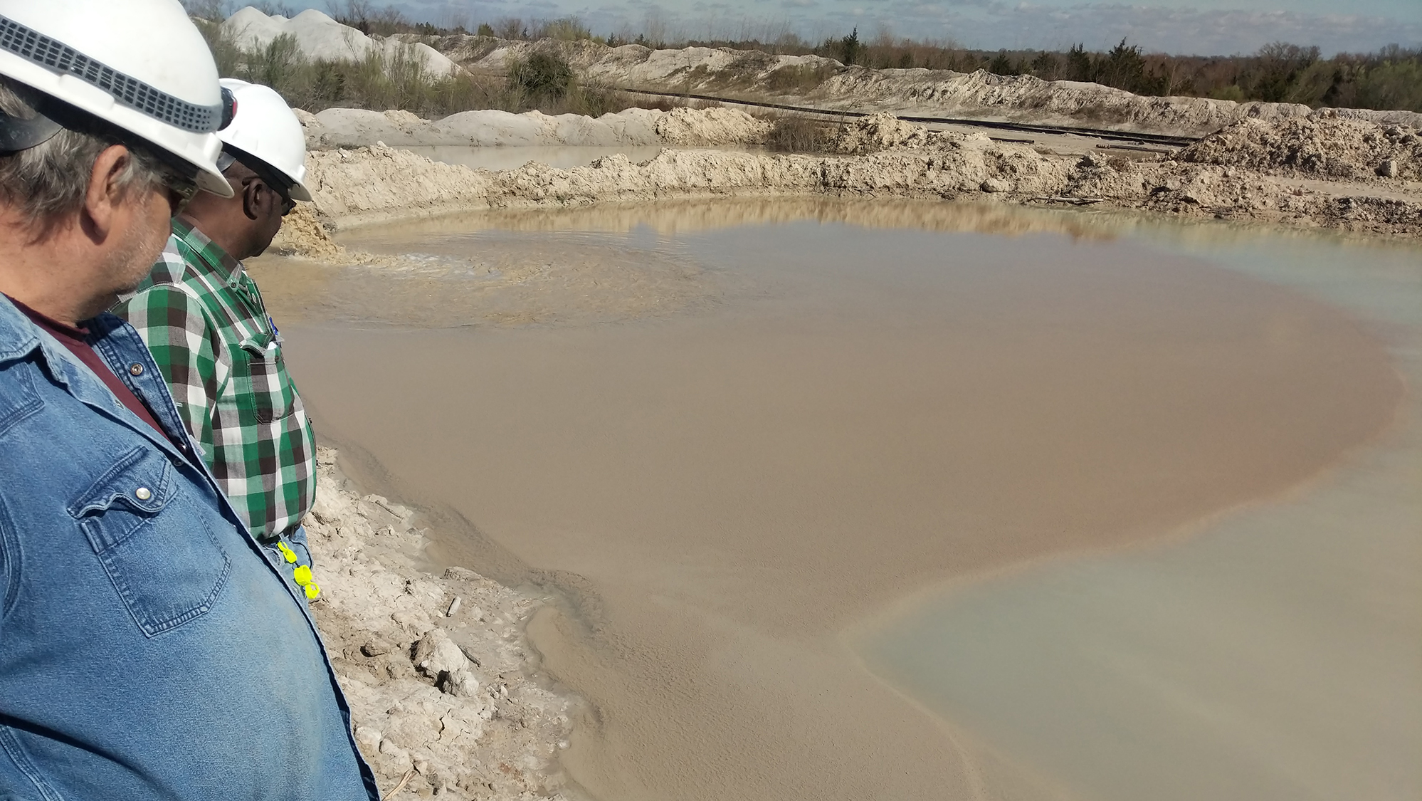 Two men in hard hats looking at dirty water discharging into a settling basin.